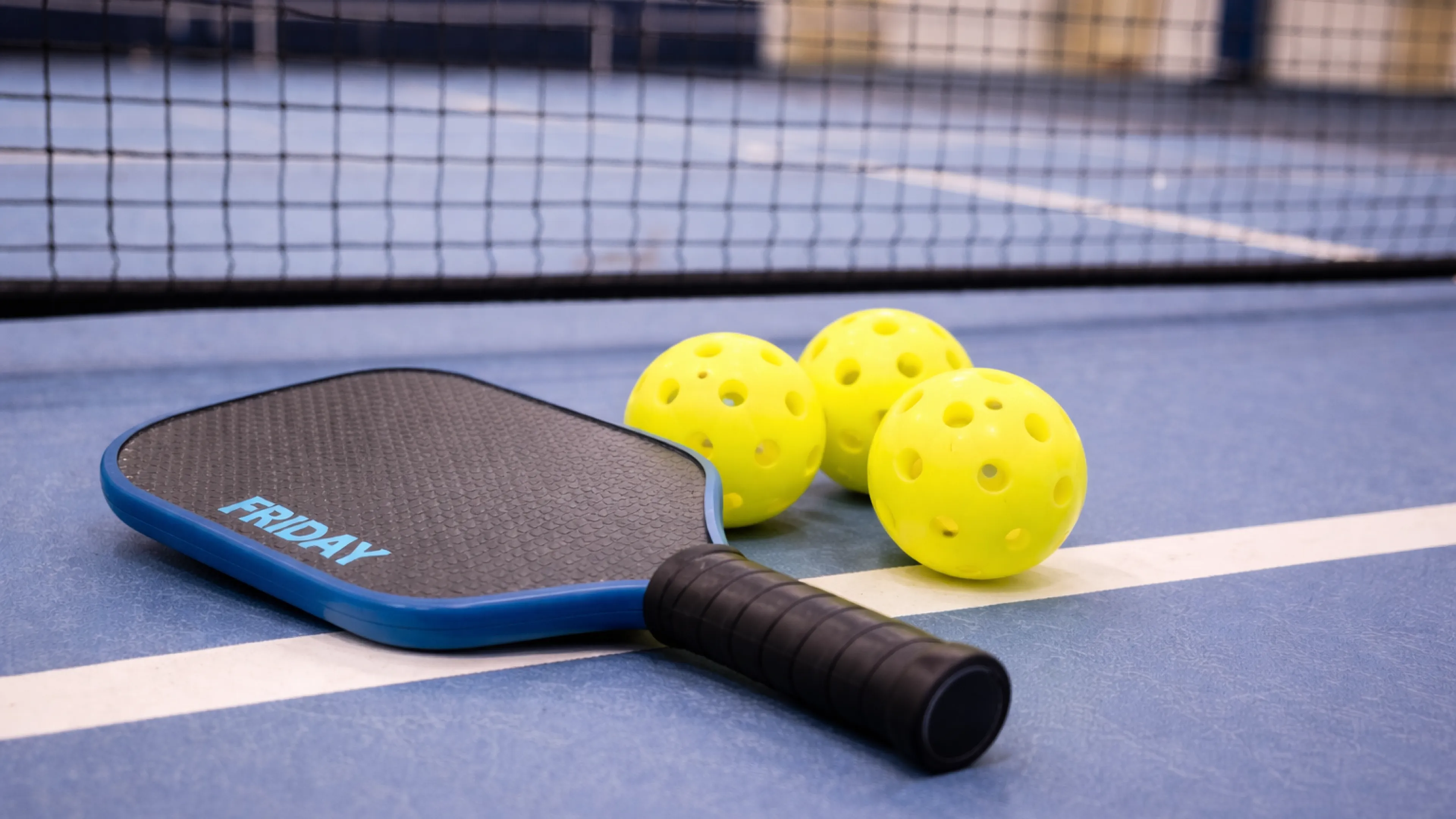Pickleball paddle and yellow pickleballs on blue court in front of net